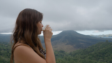 A thoughtful and happy asian girl drinking coffee against fabulous Mountain View with volcano...