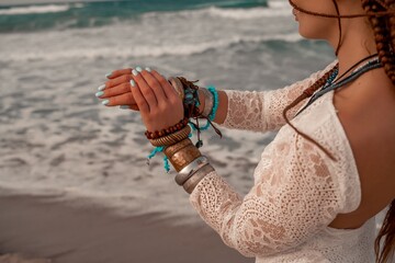 Model in boho style in a white long dress and silver jewelry on the beach. Her hair is braided, and there are many bracelets on her arms.
