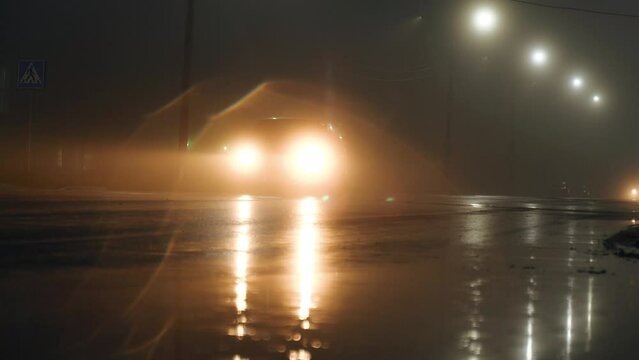 Cars with headlights on are moving along a city street at night in fog, rain. Silhouettes of cars, reflections on the pavement, blurry images.