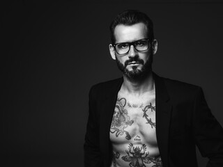 Portrait of handsome confident stylish hipster lambersexual model. Sexy modern man dressed in black jacket. Naked torso with tattoos.Fashion male posing in studio on dark background. In spectacles