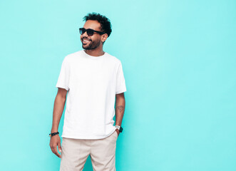 Handsome hipster smiling model. Sexy unshaven man dressed in white summer t-shirt and jeans clothes. Fashion male posing near blue wall in studio. Isolated. In sunglasses