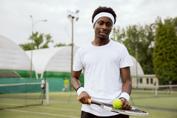 Confident tennis player stands on tennis court and poses for photo. He is holding tennis racket and ball. Guy wearing white t-shirt, headband and wristbands is looking at the camera and smiling.