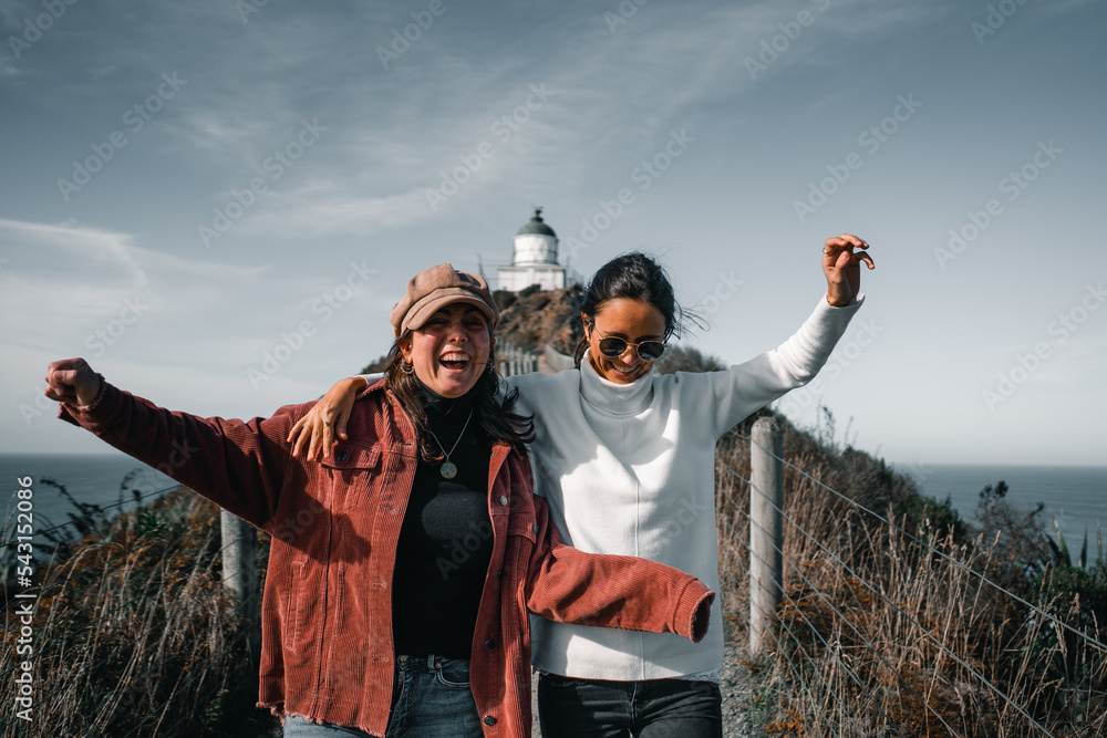 Wall mural two caucasian brunette girls hugging jumping smiling happy having fun walking on dirt road leading to lighthouse, nugget point, new zealand