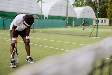 Tennis player is practicing on tennis court before tomorrow's match. He is holding racket, has head down and hands on knees. Man is wearing white T-shirt, black shorts and sneakers. He has wristbands.