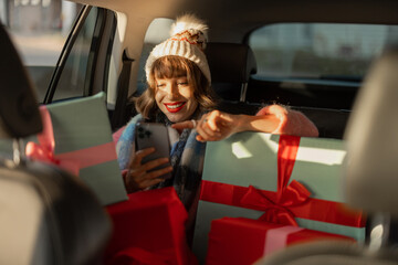 Young woman dressed for the holidays drives in car on back seat with beautifully wrapped Christmas presents and using phone. In anticipation of the winter holidays, preparation and shopping concept