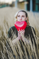 thoughtful young woman hoping or searching for self love and holding a red heart air balloon...
