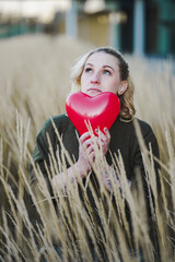 thoughtful young woman hoping or searching for self love and holding a red heart air balloon...