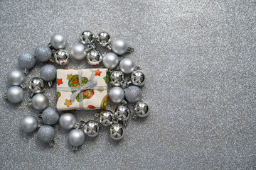 close up gift box with christmas, new year print decorations and tied bow, surrounded by silver balls baubles on grey sparkle background, festive holidays time