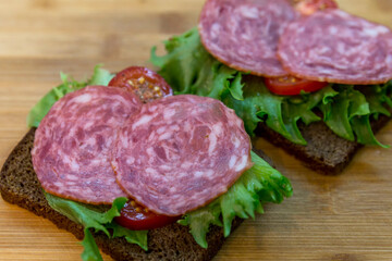 Sandwiches with sausage and herbs on a wooden board. Black bread, lettuce, tomatoes and salami. Appetizing snack. Close-up.