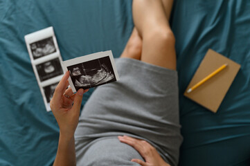 High angle shot of a pregnant woman lie down looking at a sonogram or ultrasonography picture while touching her belly in bedroom