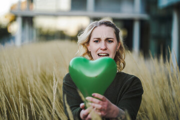thoughtful young vegan woman holding a green heart air balloon in her hand infront of her face to...