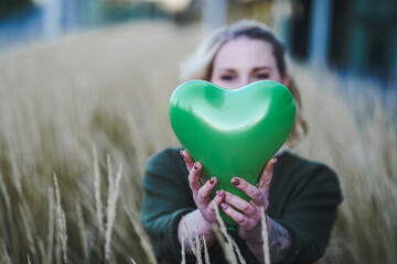 thoughtful young vegan woman holding a green heart air balloon in her hand infront of her face to...