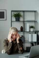 Young tired blond student or businesswoman touching head while sitting by desk in front of laptop and trying to concentrate