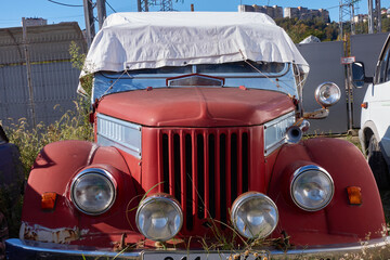 An old red car is parked in the grass. The car is covered with a white cloth from the sun. You can see chrome parts, headlight, bumper, hood, horn. Selective focus.
