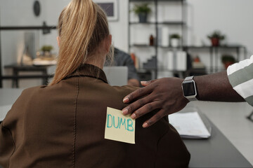 African American businessman putting sticker with word dumb on back of blond female colleague of another ethnicity while making fun of her - obrazy, fototapety, plakaty