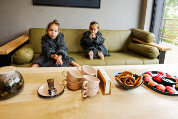 Two sisters wear bath robe sit on couch and drink tea with bagels.