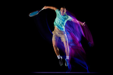 Movements in neon glow. Active male tennis player in sportwear playing tennis isolated over dark background in mixed purple neon light.