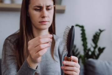 Selective focus of young girl sitting on sofa brushing hair with hair tool, worrying about hair...