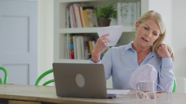 Menopausal mature woman working on laptop with connected fan at home having hot flush and fanning herself with documents - shot in slow motion