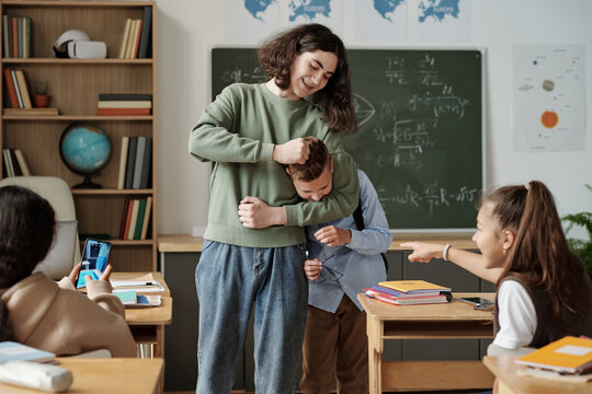 Two girls laughing at junior school learner while one of them making video of their classmate fighting with him in classroom