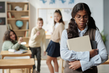 Pre-teen upset schoolgirl with books standing in front of camera with offended expression against three classmates bullying her - obrazy, fototapety, plakaty