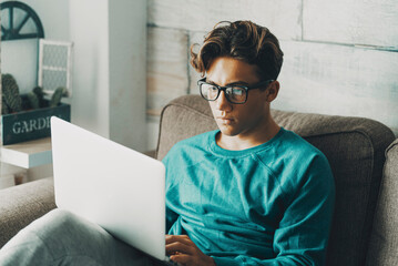 Student boy using at home a laptop sitting on the sofa with concentrated expression. Young teenager male wearing eyeglasses working on computer. Online education purpose and leisure technology