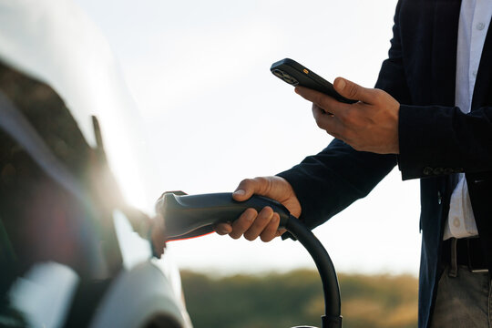 Unrecognizable businessman plugging electric car from charging station using app on smartphone. Man is plugging in power cord to an electric vehicle at sunset. Charging electric car