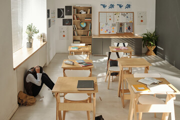 Interior of classroom and upset schoolgirl sitting on the floor between window and row of desks and...
