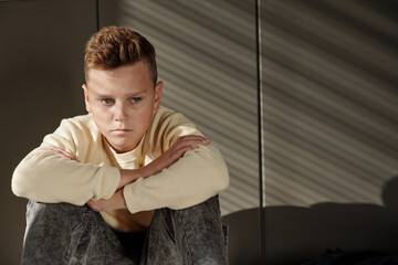 Gloomy schoolboy in casualwear looking aside while sitting on the floor after lessons and suffering...