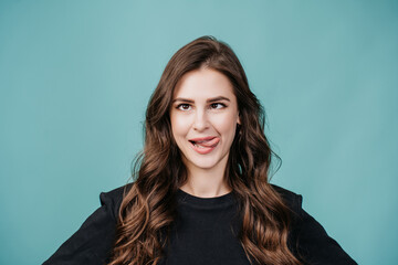 Playful brunette young woman in black sweater shows tongue, squinted eyes over turquoise background...