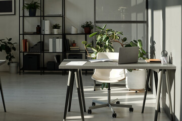 Workplace of modern manager or employee with laptop and copybooks on desk standing by sunlit wall in openspace office