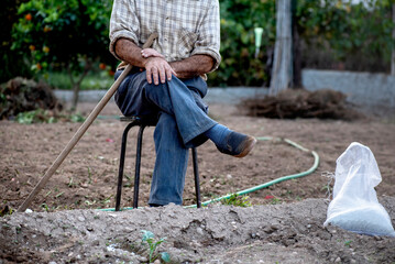 hombre blanco anciano en taburete sentado en el huerto, campesino con azada descansando de plantar, bolsa de abono a la derecha de la fotografía - obrazy, fototapety, plakaty