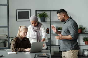 Two multicultural employees bullying young blond businesswoman sitting by desk in front of laptop...