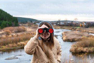 Happy young woman ecologist in beige jacket and hood looking through binoculars on the background...