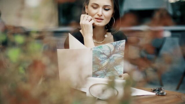 Beautiful brunette woman sitting on outdoor terrace. Caucasian female looking throught the menu in restautant. Slylish lady in bistro.