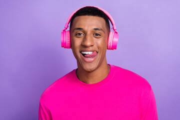 Closeup photo of young positive smiling guy stick tongue out toothy wear neon wireless beats heaphones listen stereo music isolated on violet color background