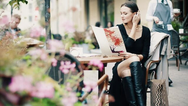 Beautiful brunette woman sitting on outdoor terrace. Caucasian female looking throught the menu in restautant. Slylish lady in bistro.