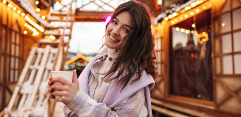 Pretty young caucsaian model looking at camera with smiling holds cup of coffee during winter day. Brunette wears shirt and sweater. Concept of rest time, lifestyle.