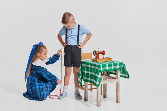 Portrait of cute little girl measuring boy's leg to sew costume isolated over grey background. Little seamstress
