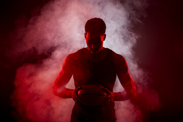 Basketball player side lit with red color holding a ball against smoke background. Serious...