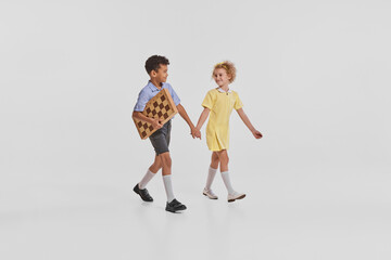 Naklejka premium Portrait of two children, boy and girl walking together to play chess isolated over grey background