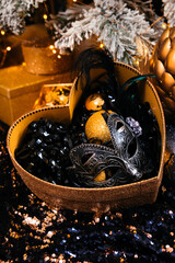 a carnival mask and Christmas decorations of gold color on black tinsel lie in a heart-shaped golden box against the background of other golden boxes, a Christmas tree garland in blur