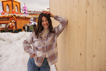 Positive young caucasian girl smiling leaned on wooden wall at amusement park. Brunette wears jeans, golf and shirt in winter. Concept of winter holidays.