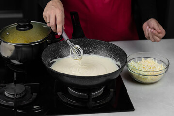 Chef cook making white cheese sauce for mac and cheese meal. Ingredients: Milk, cheese, flour and butter in wok pan on kitchen gas stove.
