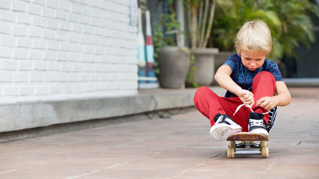 Little Skateboarder Sit On Skateboard Tying Shoelaces Before Children Training Class In Park. Active Family Lifestyle, Outdoor Activities On Summer Holidays In City. Kids Sports, Urban Fashion