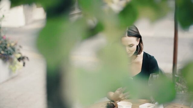 Beautiful brunette woman sitting on outdoor terrace. Caucasian female looking at the smartphone in the restautant. Slylish lady in bistro.