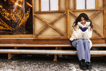 In full length cute young caucasian woman sitting near wooden house in winter day. Brunette wears casual winter clothes. Concept winter holidays.