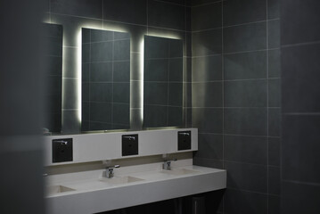 The interior of a modern public toilet, a dark-style washroom, sinks and illuminated mirrors