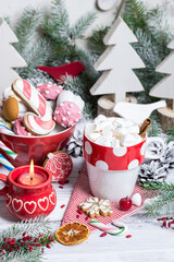 White marshmallows in cup with dots in winter composition on white wooden table decorated with natural fir tree branches, cookies, candle. Decor for Christmas holidays