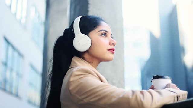 Pretty young indian businesswoman listening to music in headphones after hard work day near business centre. Young office worker with cup of coffee relaxing and looking ahead outside.